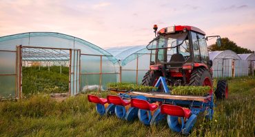 Manual seedling planter mounted to a tractor in front of greenhouses at sunset.
